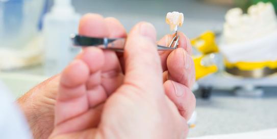 Zahntechniker hält mit einer Pinzette eine künstliche Zahnkrone in der Hand. Im Hintergrund sind unscharf Laborutensilien und Werkzeuge zu sehen.