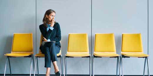 Nachdenklich wirkende Frau im Business-Outfit sitzt allein auf einer gelben Stuhlreihe in einem Wartebereich und blickt zur Seite