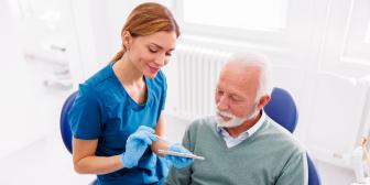 Zahnärztin in blauer Kleidung erklärt einem älteren männlichen Patienten mit grauem Haar ein Zahnmodell