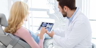 Zahnarzt im weißen Kittel zeigt einer Patientin auf einem Tablet digitale Röntgenbilder oder eine Implantatplanung