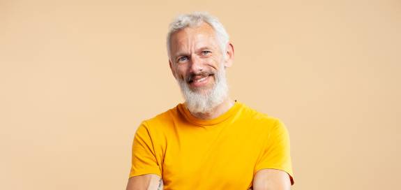Älterer Mann mit grauem Bart und Tattoos auf den Armen steht lächelnd mit verschränkten Armen vor beigefarbenem Hintergrund und trägt ein orangefarbenes T-Shirt