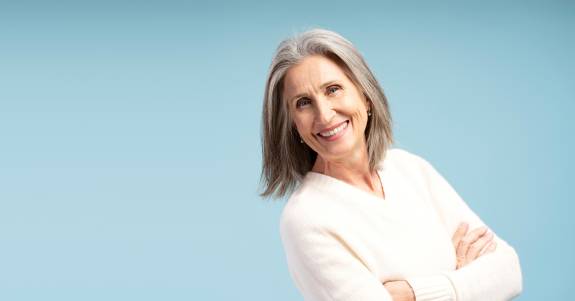 Ältere Frau mit grauen Haaren und weißem Pullover lächelt freundlich in die Kamera, mit verschränkten Armen vor hellblauem Hintergrund