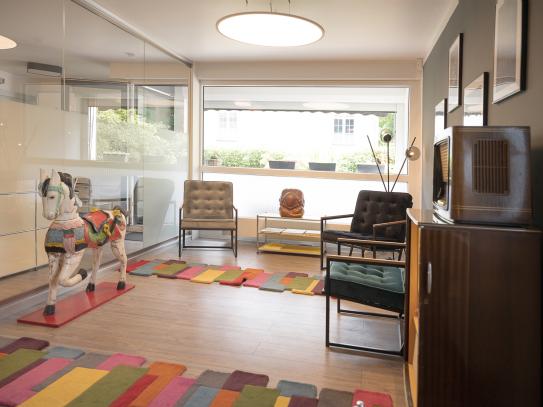 Modern eingerichtetes Wartezimmer mit bunten Teppichen, Retro-Möbeln, großen Fenstern und einem dekorativen Holzpferd.