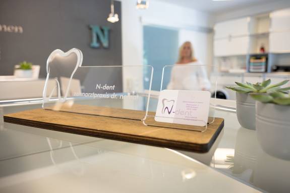 Acryl-Aufsteller mit Logo und Visitenkarte der Zahnarztpraxis „N-dent – Zahnarztpraxis Dr. Nemat“ auf einem modernen Empfangstresen.